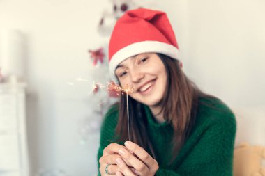 Noel Baba şapkası takan Bengal ışığıyla gülümseyen bir kız Noel ağacının arka planında bir sandalyeye oturur.