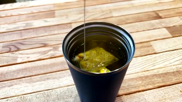 Saco de chá verde mergulhando em copo térmico preenchido com água quente na mesa de madeira — Vídeo de Stock