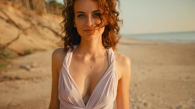 Sexy young woman with smiling face is looking at you against sunset sea background