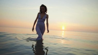 Tam vücut görünümü: Güzel ıslak kadın elbisesinin içinden size doğru yürüyor gün batımı deniz arka planında
