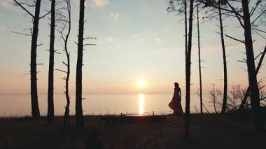 Uzun elbiseli sıska kadın gün batımına karşı yürüyor.