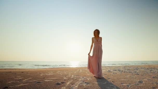 Roztomilá žena v dlouhých šatech chůze na písku na vás proti západu slunce moře pozadí — Stock video