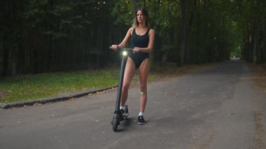 Confident young woman stands near her electric scooter wearing sneakers and a black body in the evening