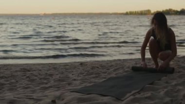 Young woman finished training and rolls away a yoga mat against a beautiful evening lake real time