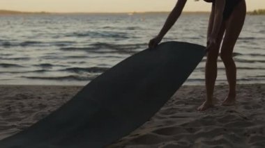Young woman prepares and unrolls a yoga mat then sits on it against a beautiful evening lake real time