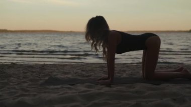 Young fit woman practices yoga wearing black body a sandy beach using a mat in the evening