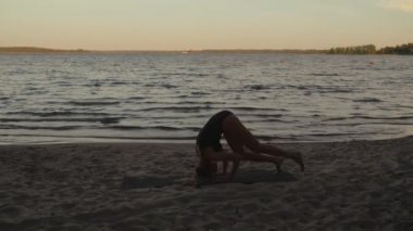Young calm woman practices yoga headstand on a lake beach wearing a black body in the evening