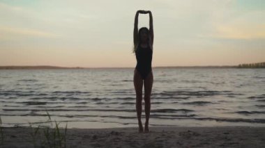 Silhouette: Fit woman practices yoga on a sandy beach against evening lake real time