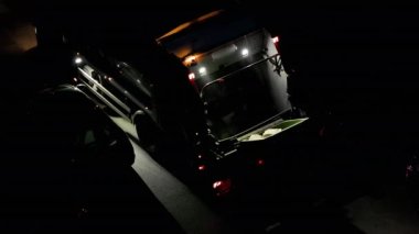 Garbage man disposes a large green bin into a garbage truck at night between many parked cars slow motion