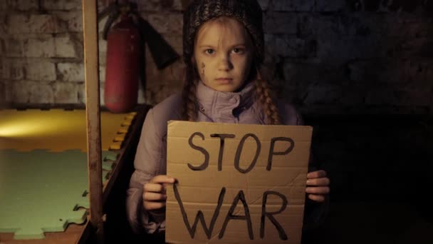 Niña ucraniana protestando por conflicto de guerra levanta pancarta con inscripción, Mensaje, texto Stop War sentado en el refugio, sótano. Crisis, paz, agresión, niño contra la guerra. — Vídeos de Stock