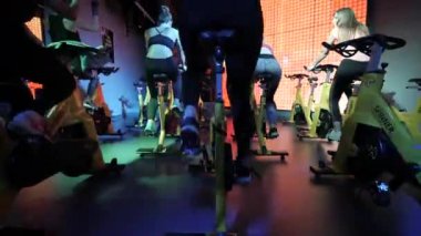 Grup sporları. Formda kadınlar. Durağan bir bisiklette antrenman. Spor salonunda spor eğitimi ekipmanları. Simülatörlerde kardiyo rutini.