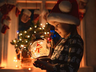 Küçük kız Noel fenerine bakıyor. Geceleri Noel Baba şapkasıyla yüzünü aydınlatıyor.,
