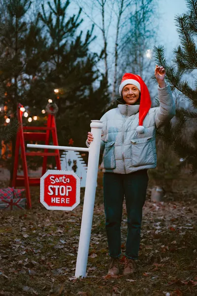 Mujer hipster con un sombrero de Santa Claus en la noche con un destello ilumina una señal de stop con el texto Santa parada aquí. — Foto de Stock