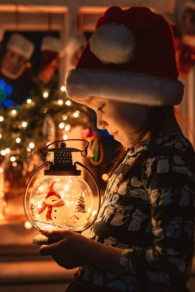 Bambina guarda una lanterna di Natale che illumina il viso in cappello Babbo Natale di notte nella stanza, Foto Stock Royalty Free