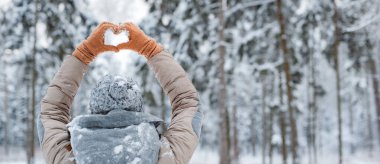 Karlı kış gününde ormanda yürüyen mutlu bir kadın. Kadın eldivenler giyiyordu. Sırtı griydi ve elleri karlı kış ormanlarında kalp hareketi yapıyordu.