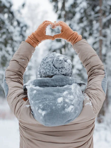 Mujer mitones vestido gris de moda se para con su espalda hace gesto del corazón con sus manos contra el fondo nevado bosque de invierno — Foto de Stock