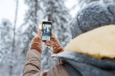 Sıcak giysili ve güneş gözlüklü mutlu bir kadın kış ormanlarında karla kaplı ağaçlara ve düşen karlara karşı akıllı telefonuyla selfie çekiyor. Soğuk bir günde kadın kendi portresini çekiyor.
