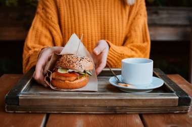 Sağlıklı fast food. Turuncu kazaklı, elinde lezzetli görünen hamburger tutan, susamlı, sebzeli, yeşillikli, tavuklu ve çedar peynirli bilinmeyen bir kızın kesilmiş fotoğrafı. Kapat.