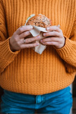 Sağlıklı fast food. Turuncu kazaklı, elinde lezzetli görünen hamburger tutan, susamlı, sebzeli, yeşillikli, tavuklu ve çedar peynirli bilinmeyen bir kızın kesilmiş fotoğrafı. Kapat.