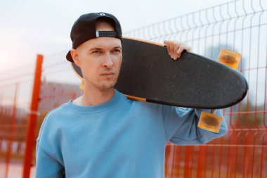 Şık şapkalı, tahta kaykaylı genç bir adamın portresi. Omuzunda tahta bir kaykayla dikiliyor, dikkatlice başka yere bakıyor ve şehir parkında aktif olarak vakit geçiriyor. Kaykay sporu konsepti