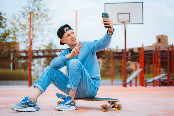 Full-leghth of young hipster guy in cap makes selfie with mobile phone while sitting on skateboard, gestures victory sign with fingers, skateboarder sharing photos with followers on social networks