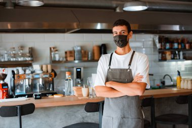 Kendine güvenen genç bir kahve dükkanı sahibi ya da yüz maskesi ve önlüklü bir barista kollarını kavuşturmuş, son moda kafenin ahşap tezgahının önünde poz veriyor covid-19 pandemisi sırasında. Yeni normal kavram