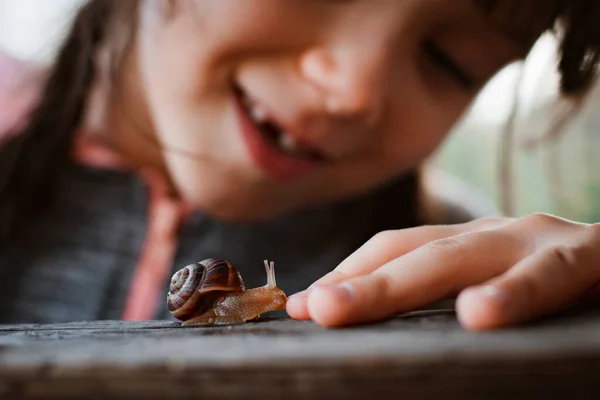 Kleines Mädchen beobachtet aufmerksam kleine Schnecke, die auf Holzbank kriecht, während sie Zeit in der Natur verbringt lizenzfreie Stockbilder