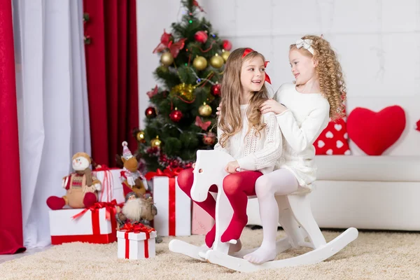 Christmas, x-mas, winter, happiness concept - two adorable happy girls playing on horse near the christmas tree — Stock Photo, Image