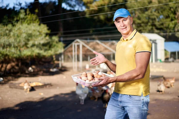 Orgulhoso jovem segurando uma cesta cheia de ovos com galinhas atrás dele . — Fotografia de Stock