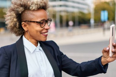 Kıvırcık saçlı, açık havada mutlu gülümseyen akıllı telefon kullanan Afro-Amerikan kadın.