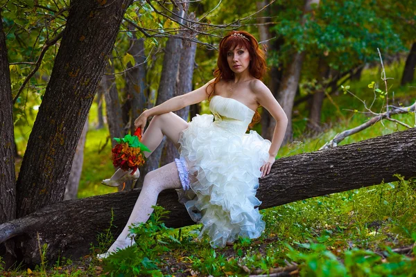 Rothaarige Braut im weißen Kleid mit einem Strauß auf einem Baum sitzend — Stockfoto