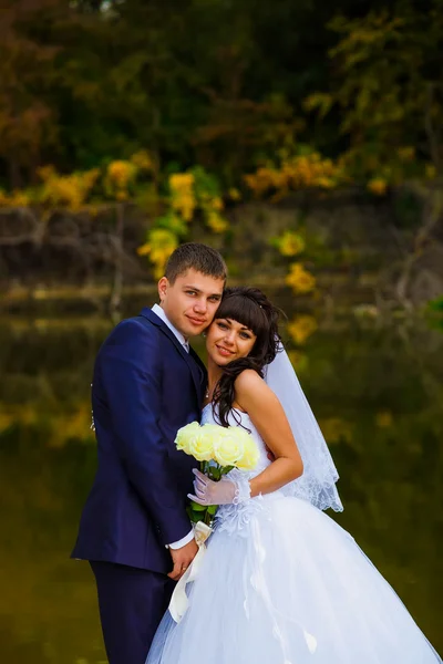 Bride and groom standing at the water behind the yellow leaves f — Stock Photo, Image