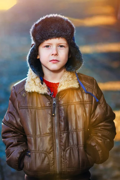 Sunlight boy in brown jacket and fur hat on street on blue abstr — Stock Photo, Image