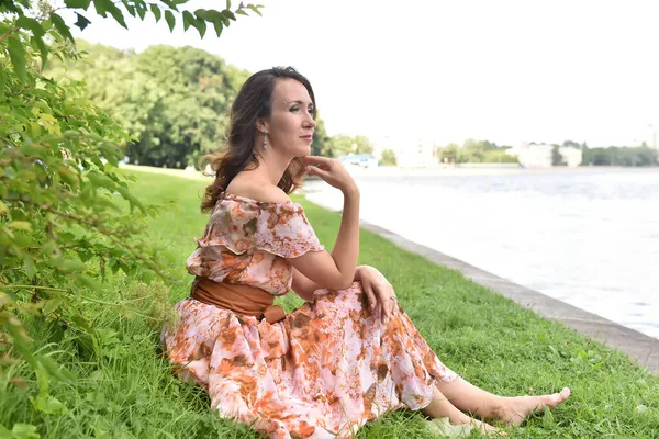 穿着粉色衣服的漂亮的黑发女人坐在河边的草地上 — 图库照片