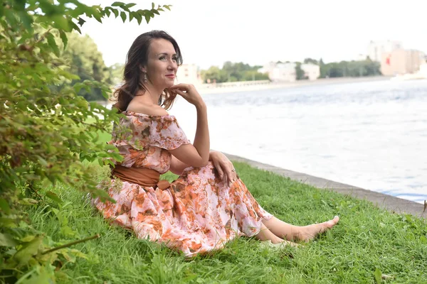 穿着粉色衣服的漂亮的黑发女人坐在河边的草地上 — 图库照片