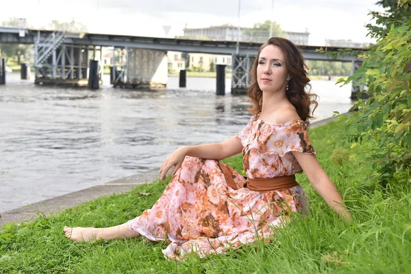 ピンクのドレスを着た美しいブルネットの女性が川のそばの草の上に座り — ストック写真