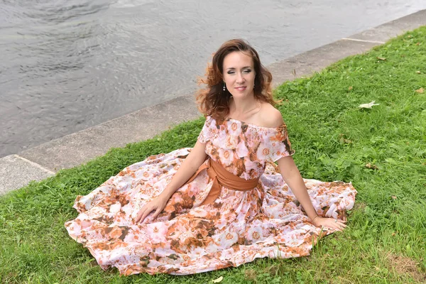 ピンクのドレスを着た美しいブルネットの女性が川のそばの草の上に座り — ストック写真