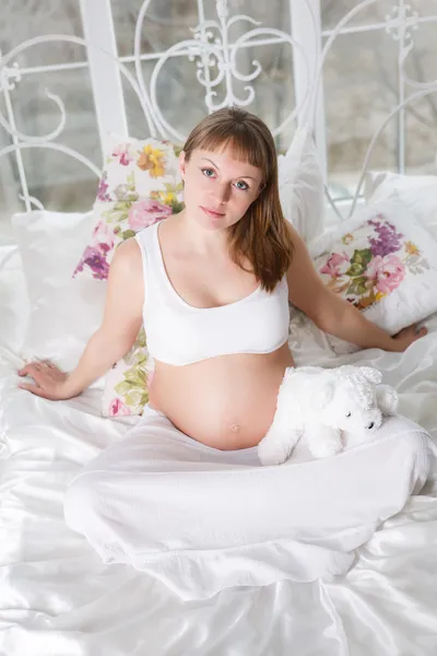 Belle femme enceinte avec des bottines pour enfants posant tout en s'asseyant sur un lit — Photo