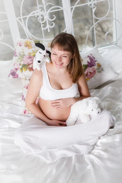 Hermosa mujer embarazada con los botines de los niños posando mientras se sienta en una cama —  Fotos de Stock