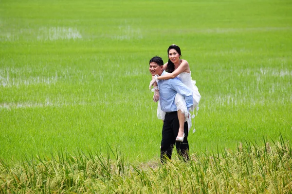 Smiling groom carrying on his back bride in paddy field — Stock Photo, Image
