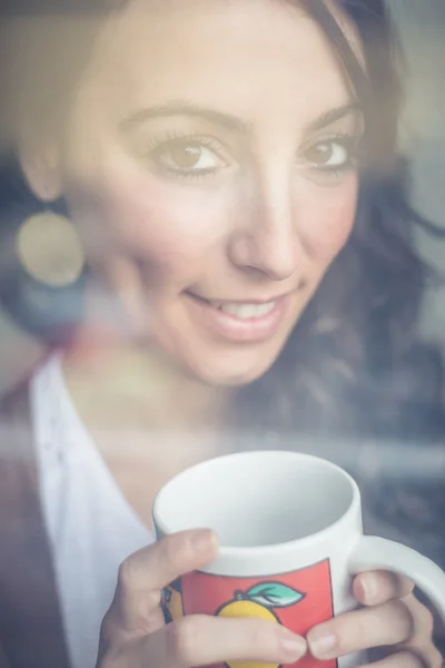 Schöne Frau mit Tasse, die durch Fenster blickt — Stockfoto