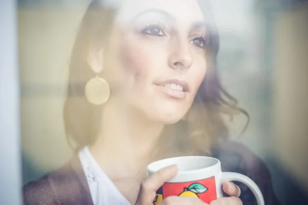 Schöne Frau mit Tasse, die durch Fenster blickt — Stockfoto