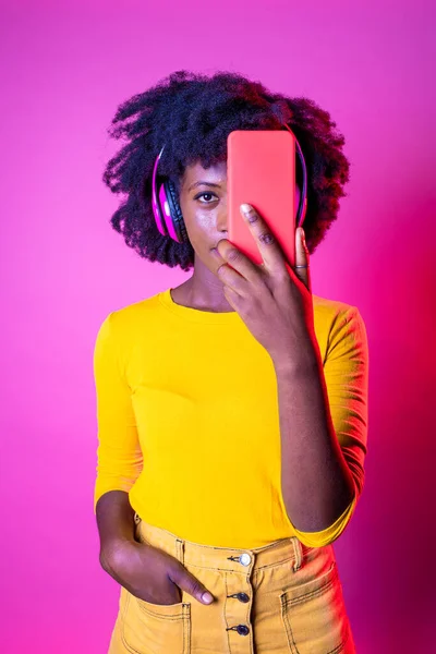 若い黒の女性は 無線技術のヘッドフォンを装着スマートフォンで彼女の顔をカバー孤立した背景 — ストック写真