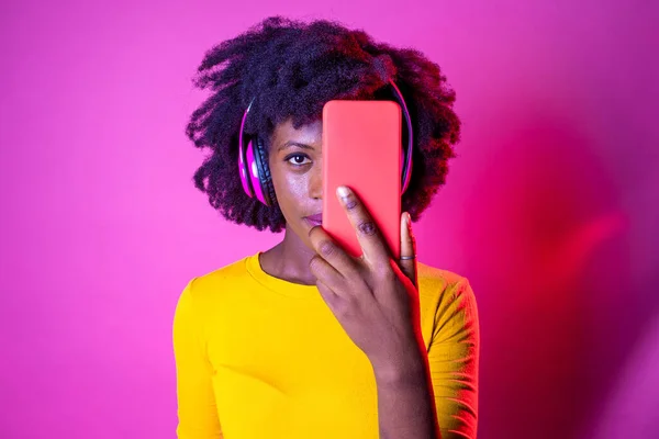 若い黒の女性は 無線技術のヘッドフォンを装着スマートフォンで彼女の顔をカバー孤立した背景 — ストック写真