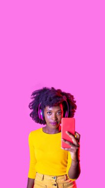 Akıllı telefon tutan genç siyahi kadın selfie çekiyor ya da kablosuz kulaklık takıyor. Reklam alanının arka planında izole edilmiş.