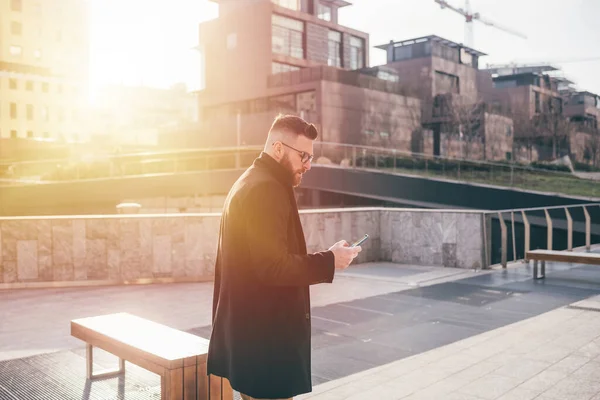 若い髭の白人ビジネスマン屋外バックライト街の夕日スマートフォンを使って屋外を歩く — ストック写真