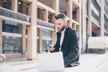 Genç, sakallı, beyaz, çağdaş iş kumandalı, açık havada esnek, akıllı telefon ve bilgisayar kullanan bir işçi.