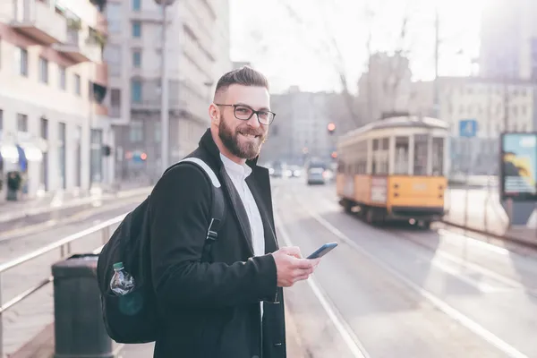 スマートフォンを使ってバスステーションで現代的な髭を生やしたビジネスマンの笑顔肯定的な笑顔 — ストック写真