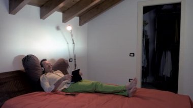 Yetişkin bir adam evde yatarken, tablet video kullanarak kulaklık takıyor. İletişim, sohbet, teknoloji konsepti.