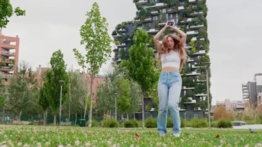 Genç beyaz kadın kendini özgür hissediyor ve elinde akıllı telefonla dans ederek eğleniyor.